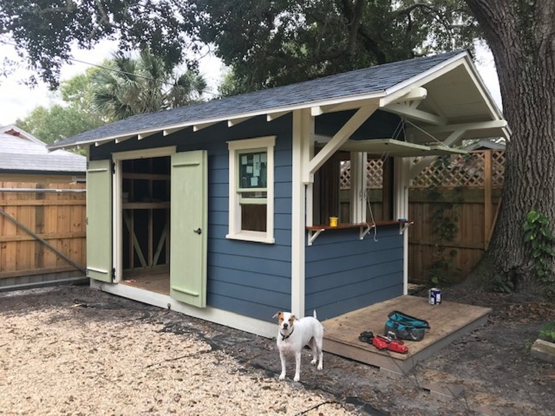 Photo of grey-blue shed with double side doors and a serving window with bar. Back yard setting with gravel and large overhanging trees.
